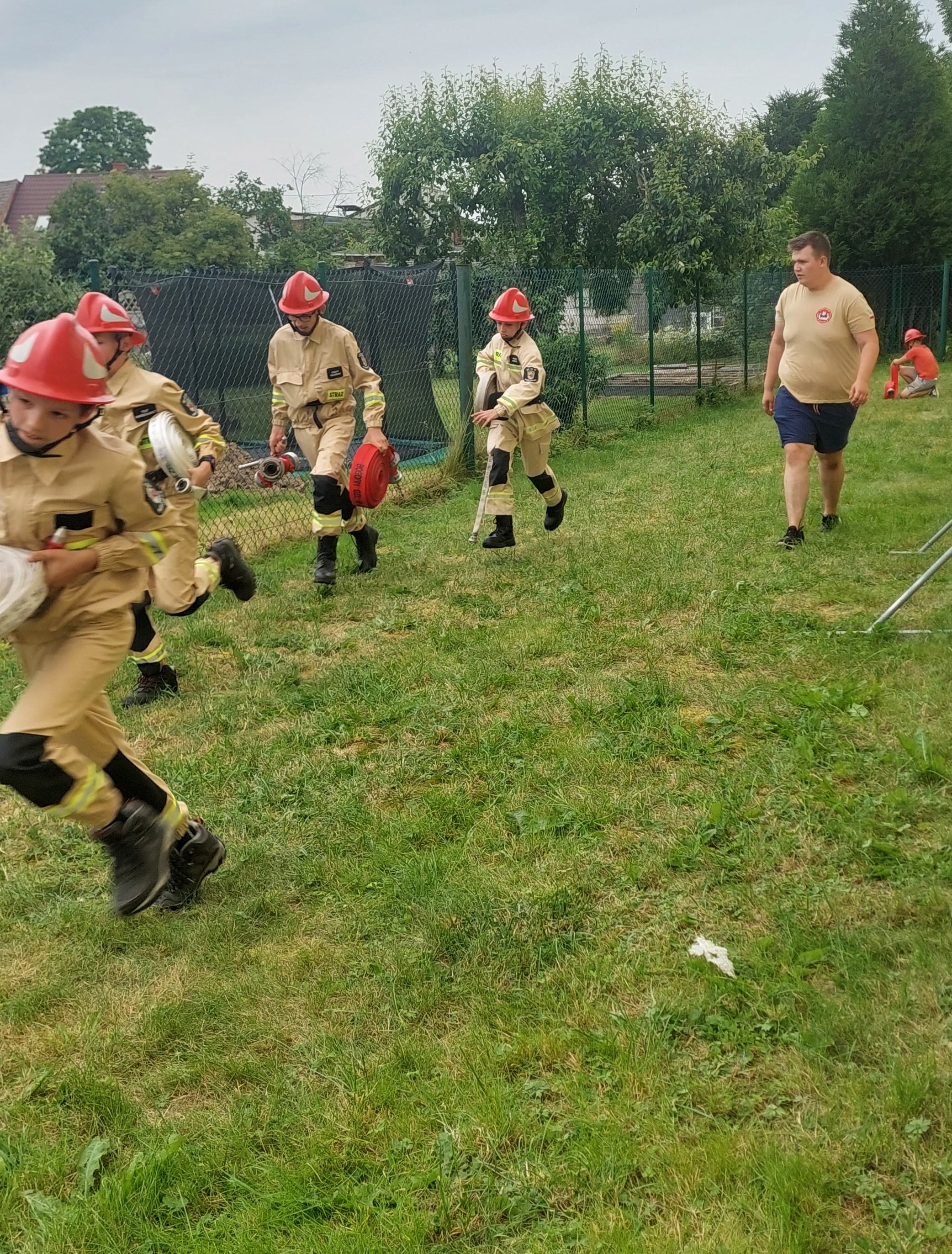 Szkolenie do biegu bojowego Członkowie Mżodzieżowej Drużyny Pożarniczej w biegu ze zwiniętymi wężami, w tle instruktor