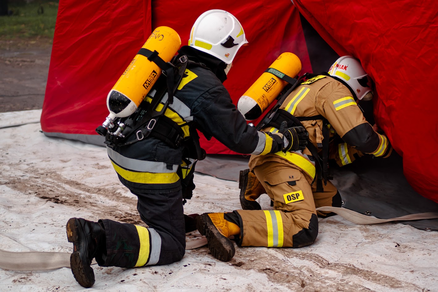 Wejscie do komory dymowej w asyście prowadzącego szkolenie Instruktor pomaga strażakowi wejść do komory dymowej podczas ćwiczeń