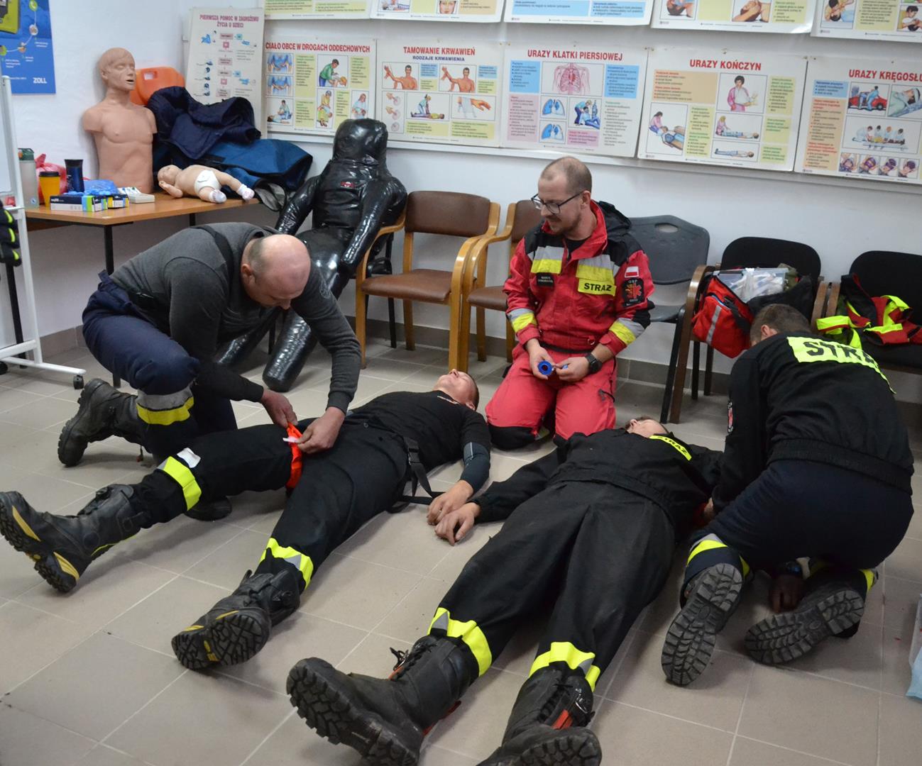 Zajęcia z pierwszej pomocy - tamowanie krwotoków opaską uciskową Dwóch strażaków udziela pierwszej pomocy poszkodowanym kolegom przez założenie opasek uciskowych, w środku - instruktor