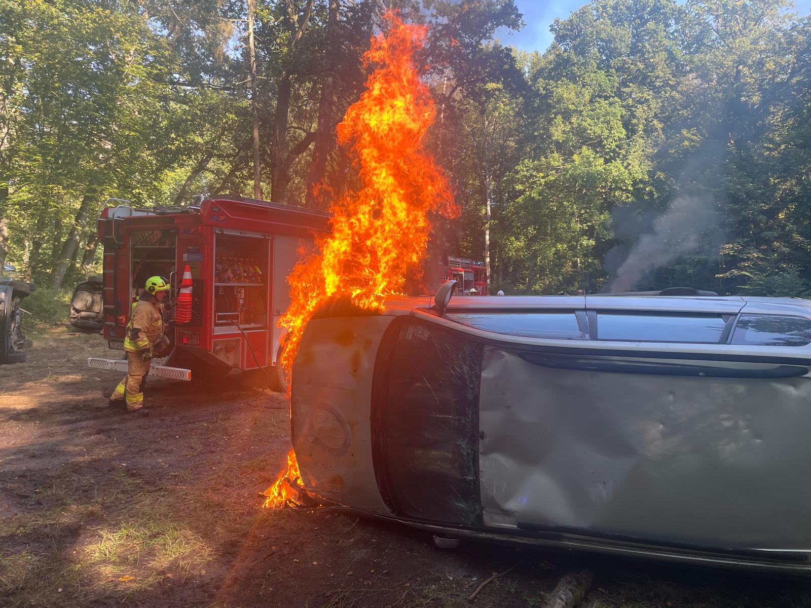 Wypadek samochodowy z pożarem pojazdu Przewrócony, płonący smaochód, w tle po lewej widoczny wóz gaśniczy straży pożarnej