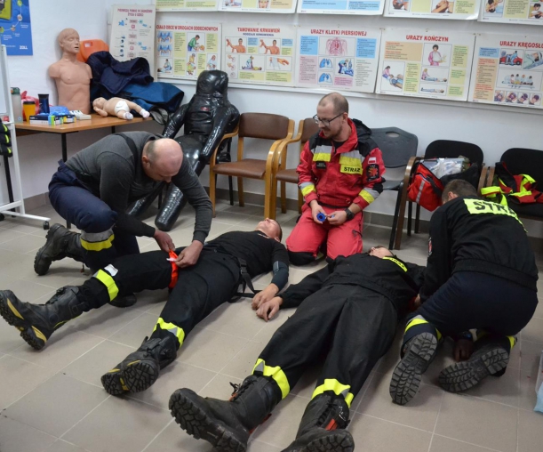 Dwóch strażaków udziela pierwszej pomocy poszkodowanym kolegom przez założenie opasek uciskowych, w środku - instruktor