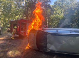 Przewrócony, płonący smaochód, w tle po lewej widoczny wóz gaśniczy straży pożarnej